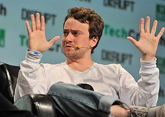 Portrait of George Hotz, a young man with short dark hair, wearing a black t-shirt, looking directly at the camera.